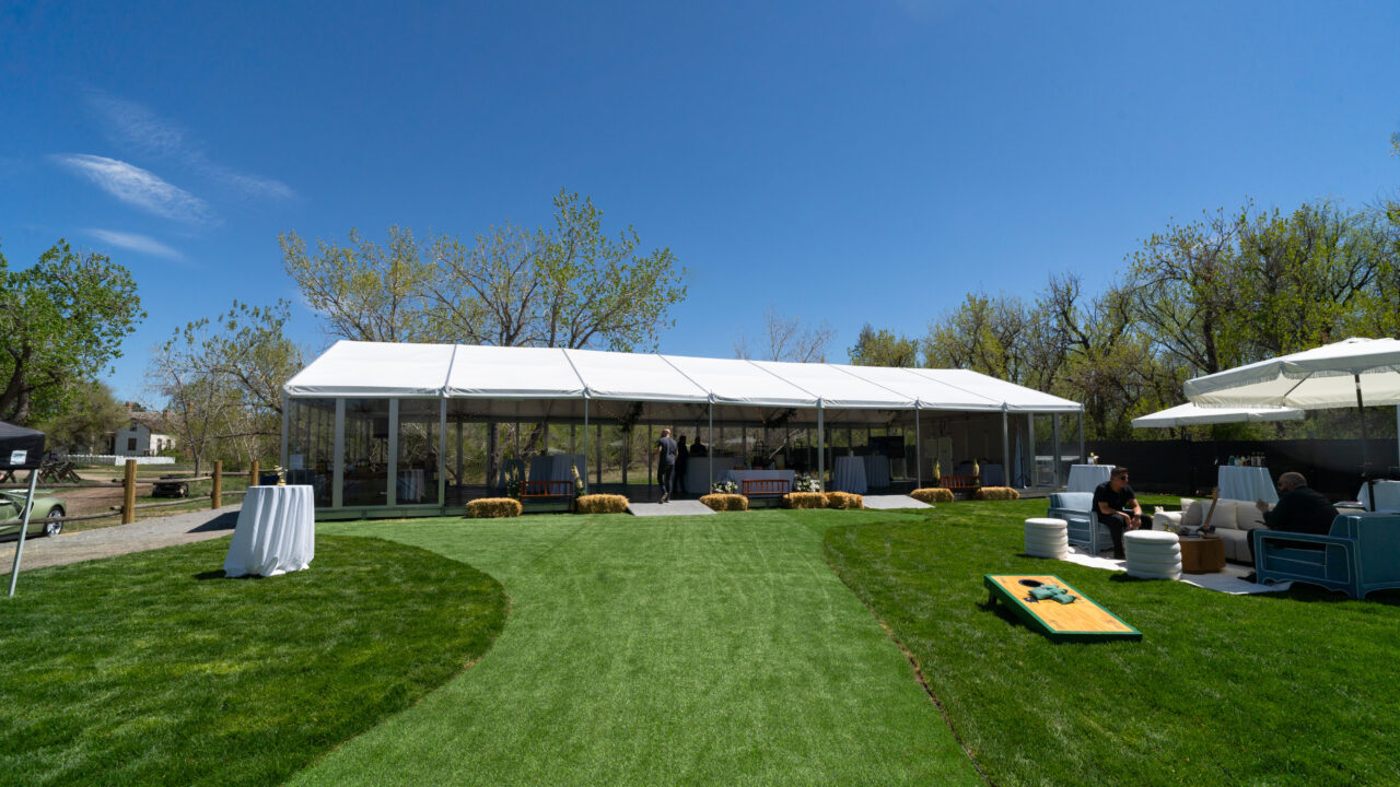 Outdoor event setup with a large white tent, hay bales, and lounge seating on a green lawn under a clear blue sky
