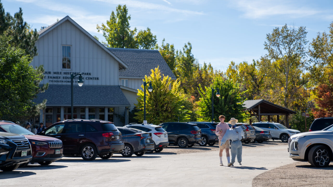 Two people walking in parking lot near Four Mile Historic Park Family Education Center, surrounded by parked cars and trees