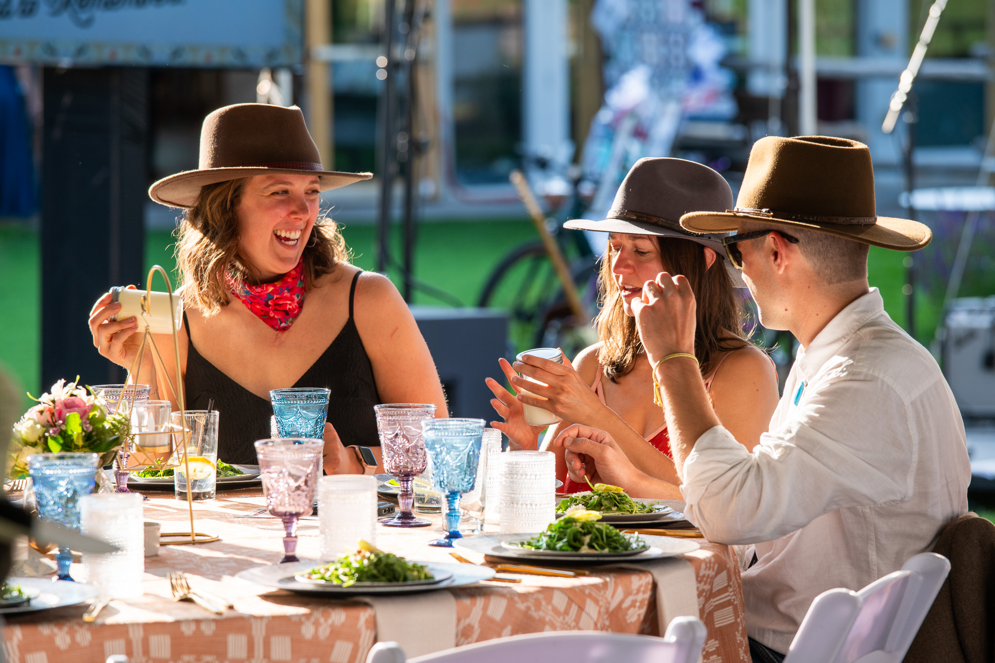 Three people in hats enjoying a sunny outdoor meal, laughing