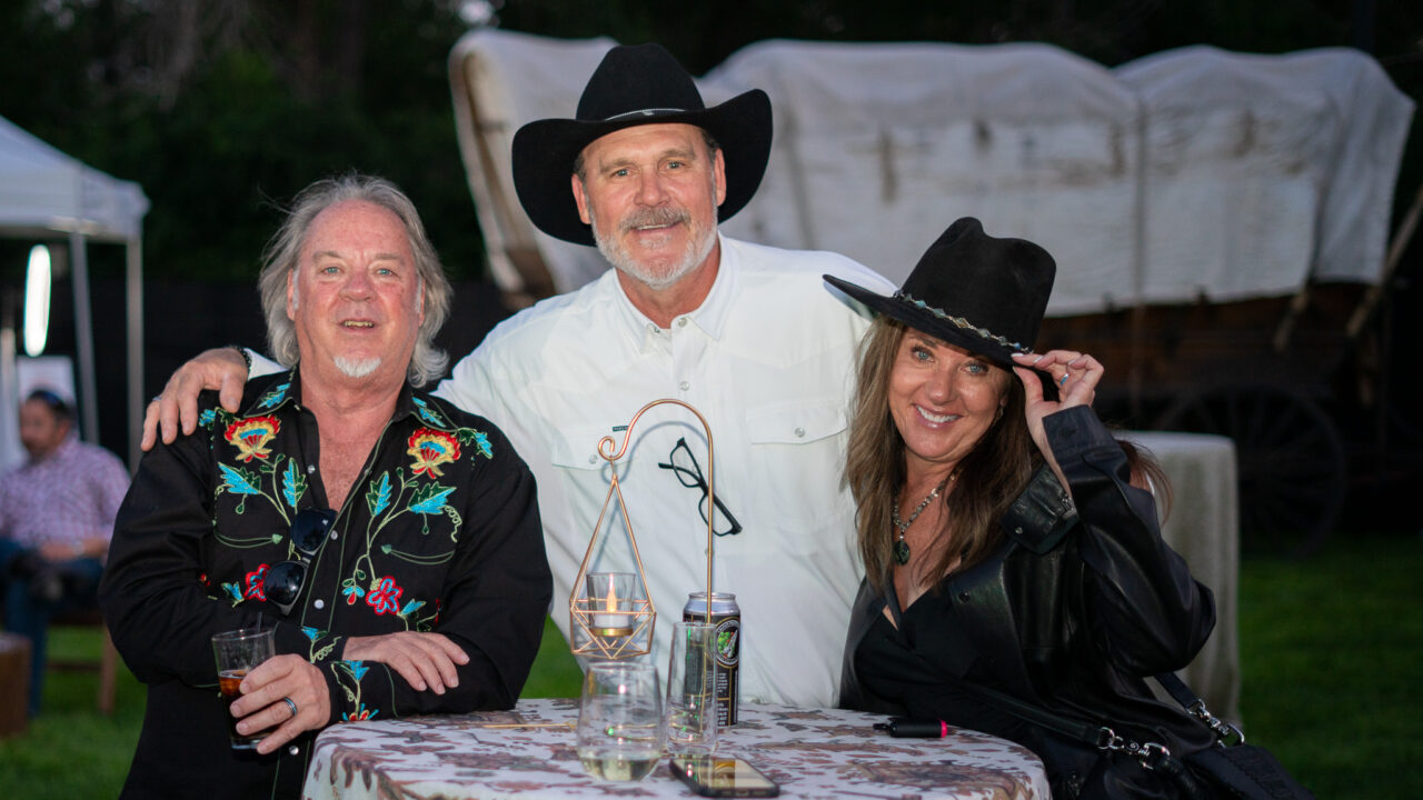 Tres personas con sombreros de vaquero sonriendo en un evento al aire libre, con una mesa y una carreta cubierta al fondo