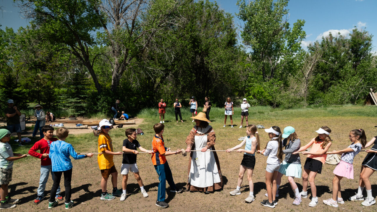 Kids and an adult in a historical costume play tug of war outdoors, surrounded by trees and onlookers