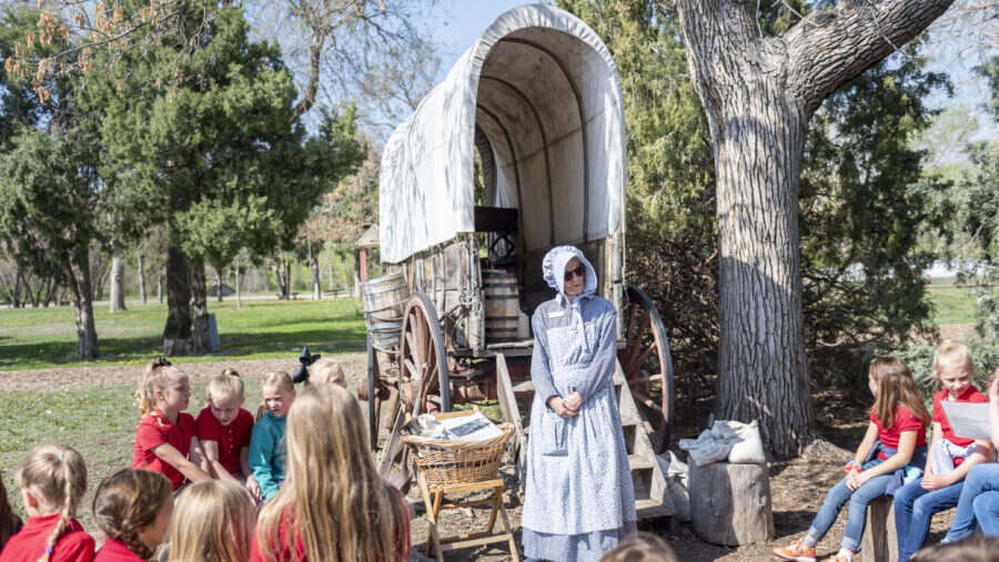 Children gather around a covered wagon, listening to a woman in period clothing, WH00017-900x670-c-aspect-ratio-640-360.jpg, explaining history