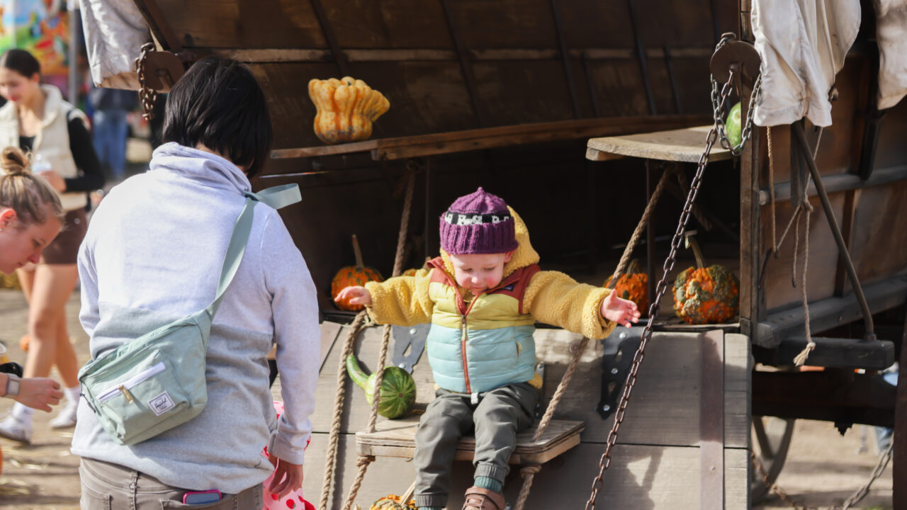 Child in colorful winter clothes sitting on a swing attached to a rustic wagon, surrounded by pumpkins, interacting with nearby people