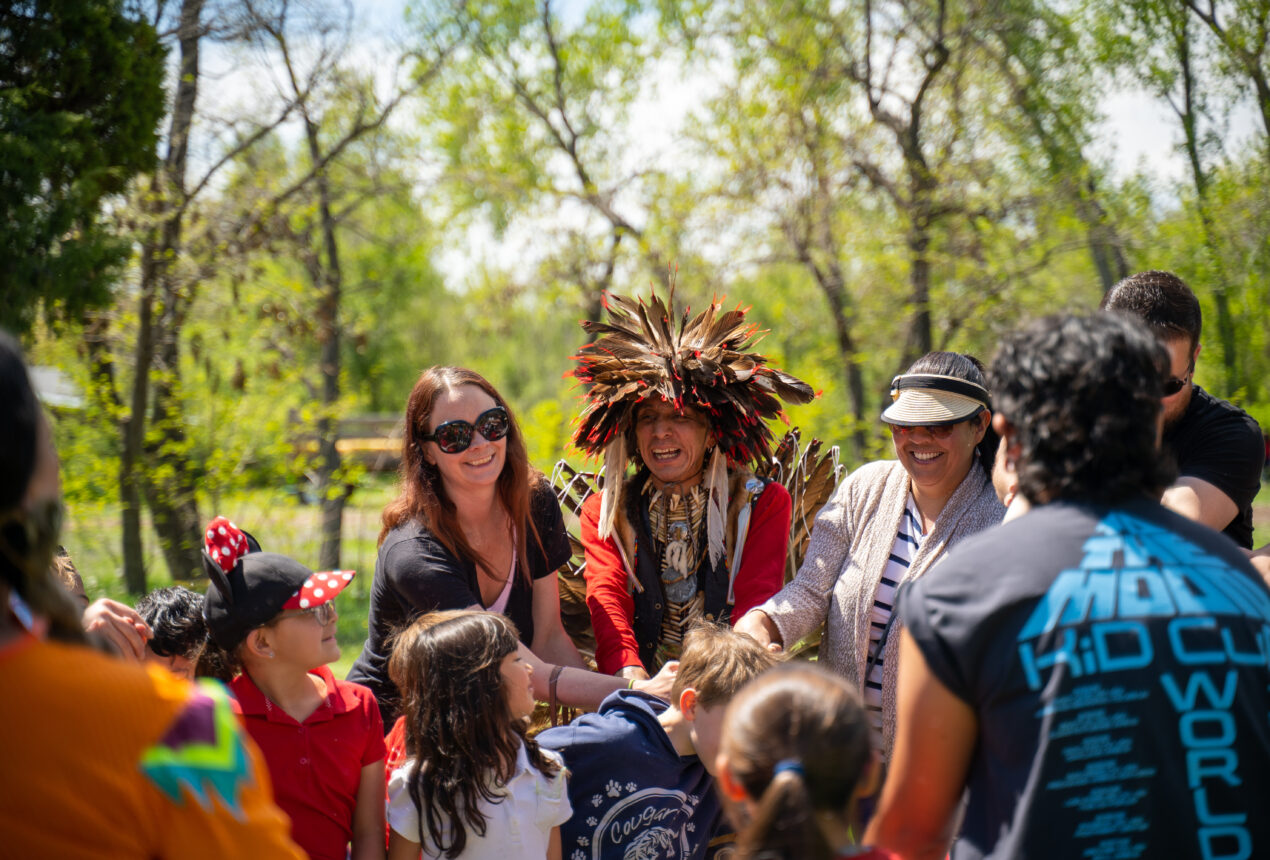 Un grupo en Four Mile Park rodea a una persona con un tocado de plumas; todos sonríen y interactúan en un entorno al aire libre