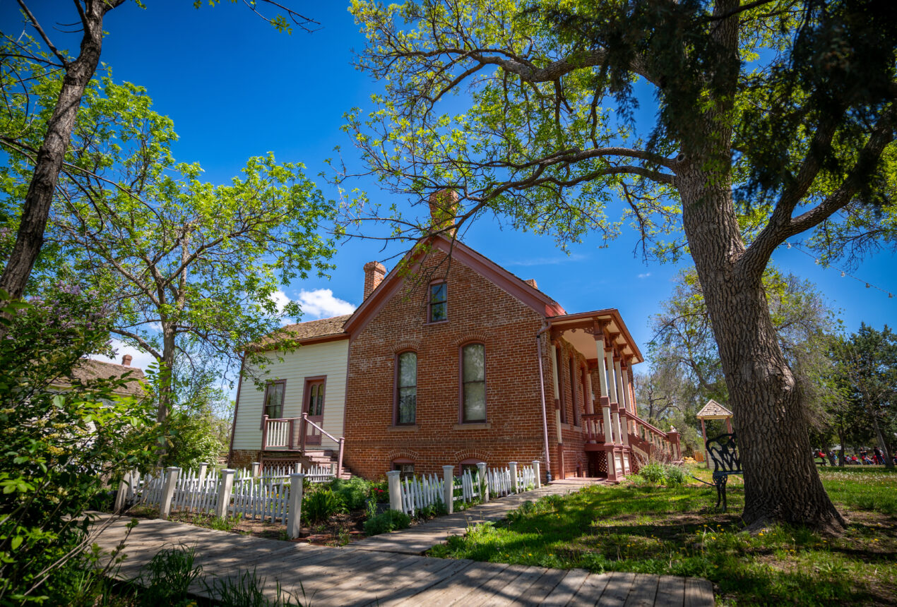 Casa histórica en un parque, con fachada de ladrillo y revestimiento de madera, rodeada de una exuberante vegetación y bajo un cielo azul brillante