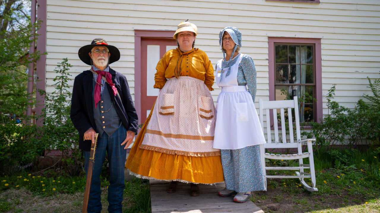 Three people in historical attire stand outside a white house