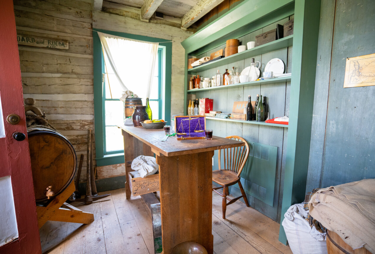Habitación rústica de estilo antiguo con muebles de madera, estanterías llenas de botellas y un cartel que dice «Alojamiento y comida»