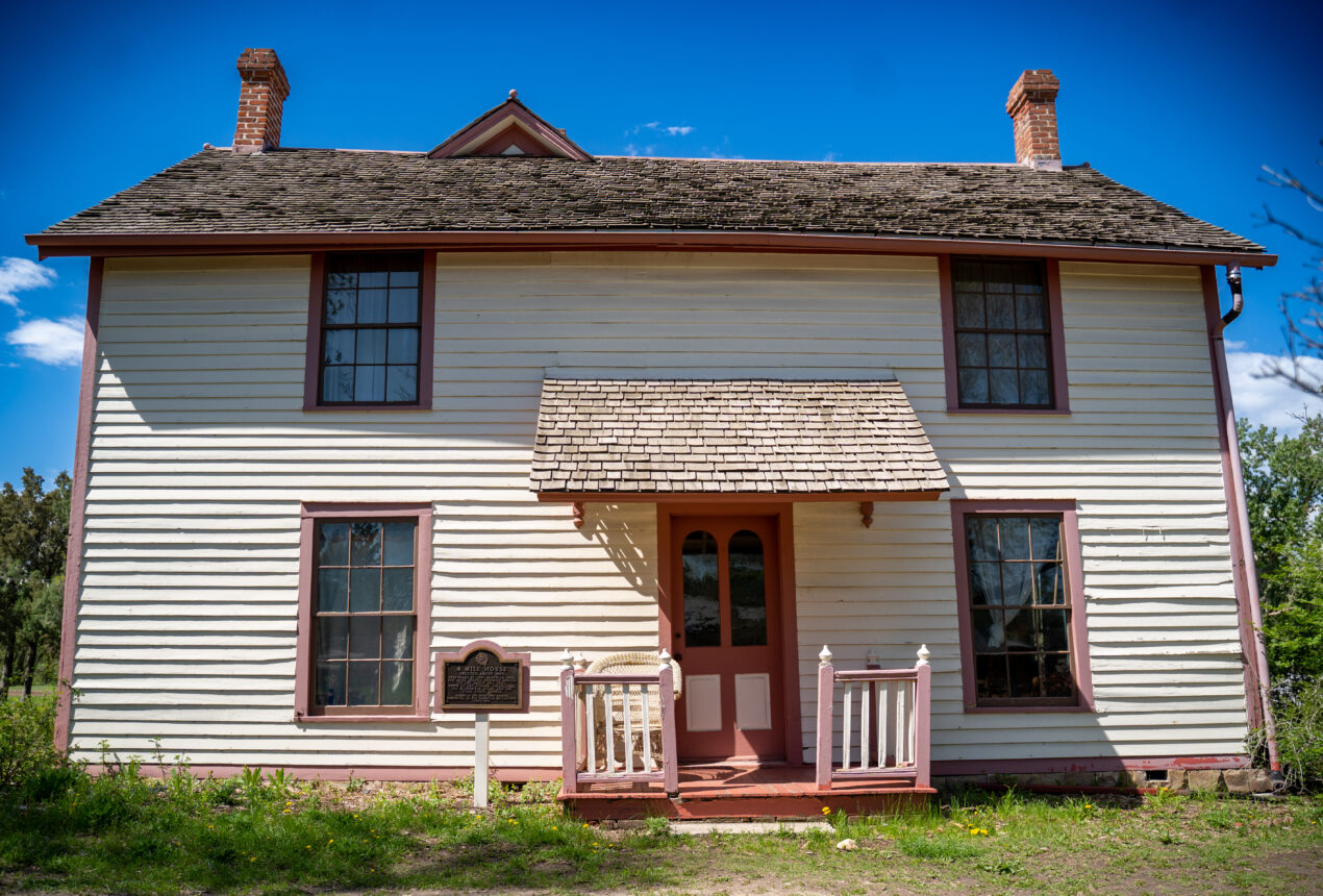 Casa histórica de dos plantas con revestimiento de madera, molduras rojas y una placa cerca de la entrada, situada en un entorno ajardinado