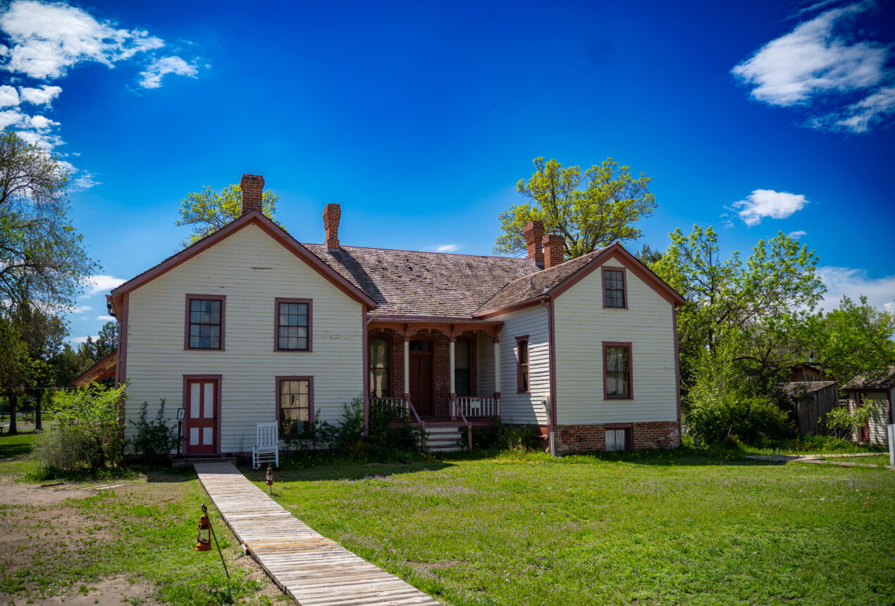 Casa histórica blanca con molduras rojas, chimenea de ladrillo y pasarela de madera, rodeada de césped verde y árboles bajo un cielo azul