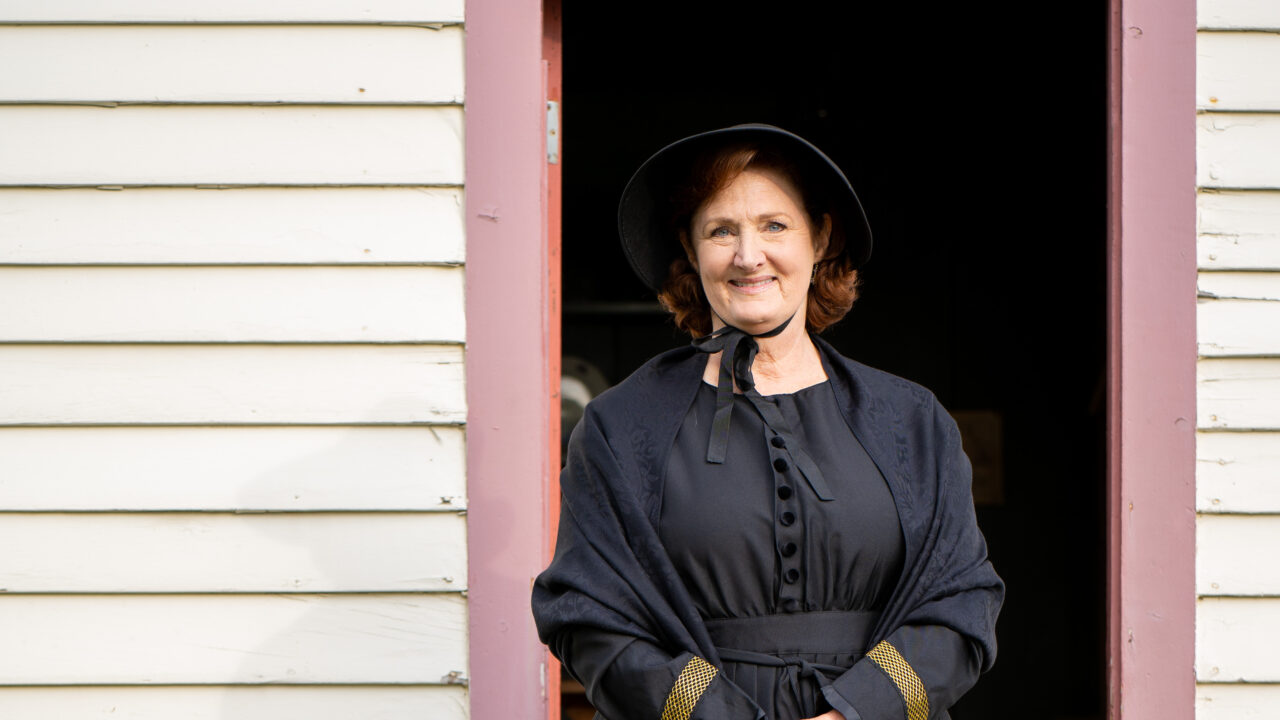 Woman in black dress and bonnet smiling, standing in doorway of wooden building
