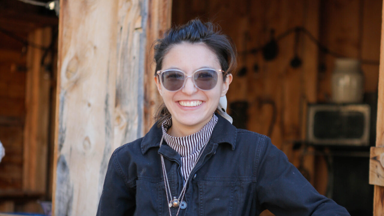 Person smiling outdoors wearing sunglasses and a striped shirt under a dark jacket