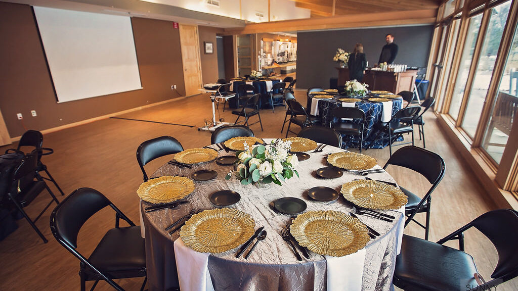 Elegant banquet room set for event with round tables, gold plate chargers, black chairs, and floral centerpieces