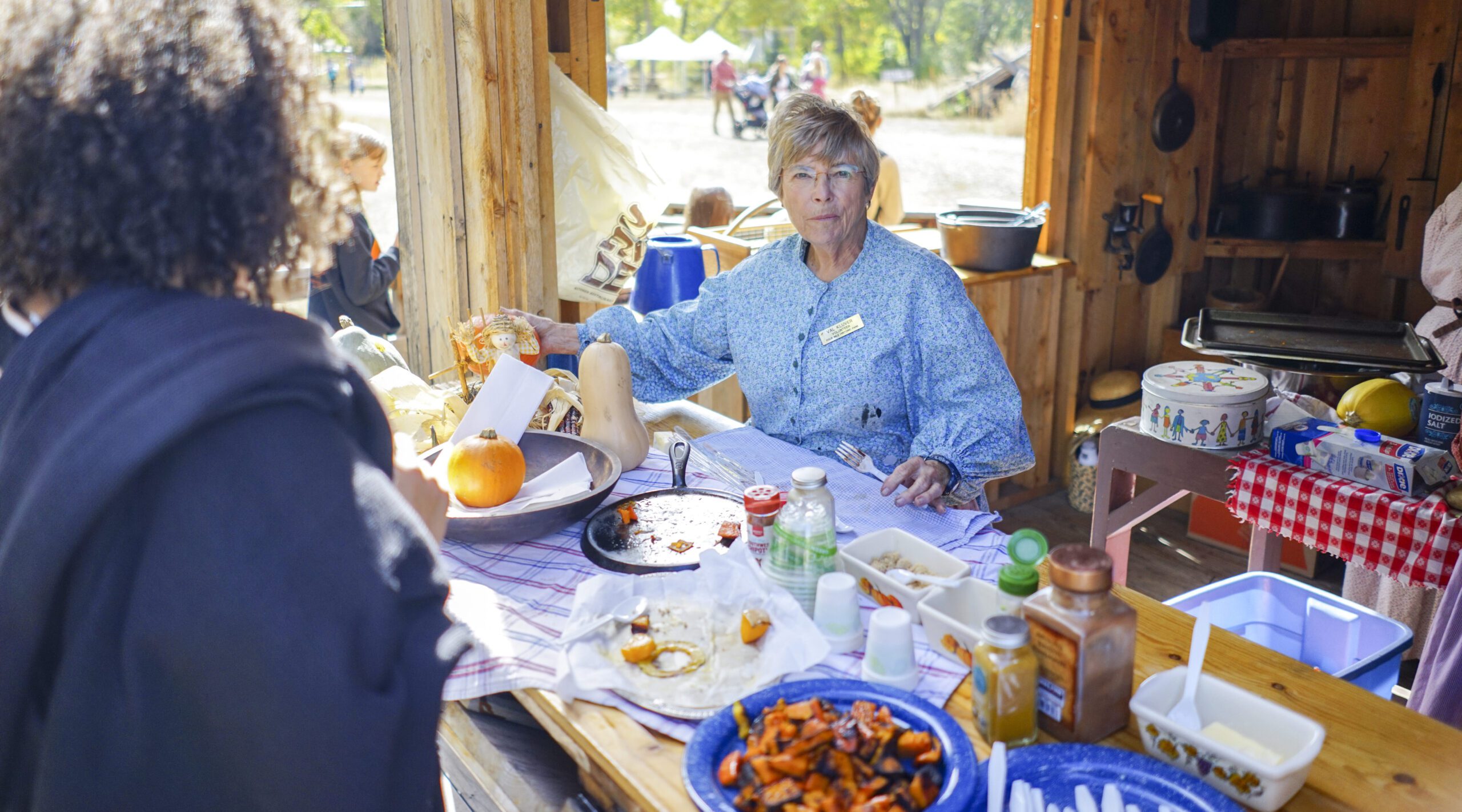 Persona vestida con ropa de época en un puesto de comida al aire libre de estilo rústico, con calabazas y utensilios de cocina
