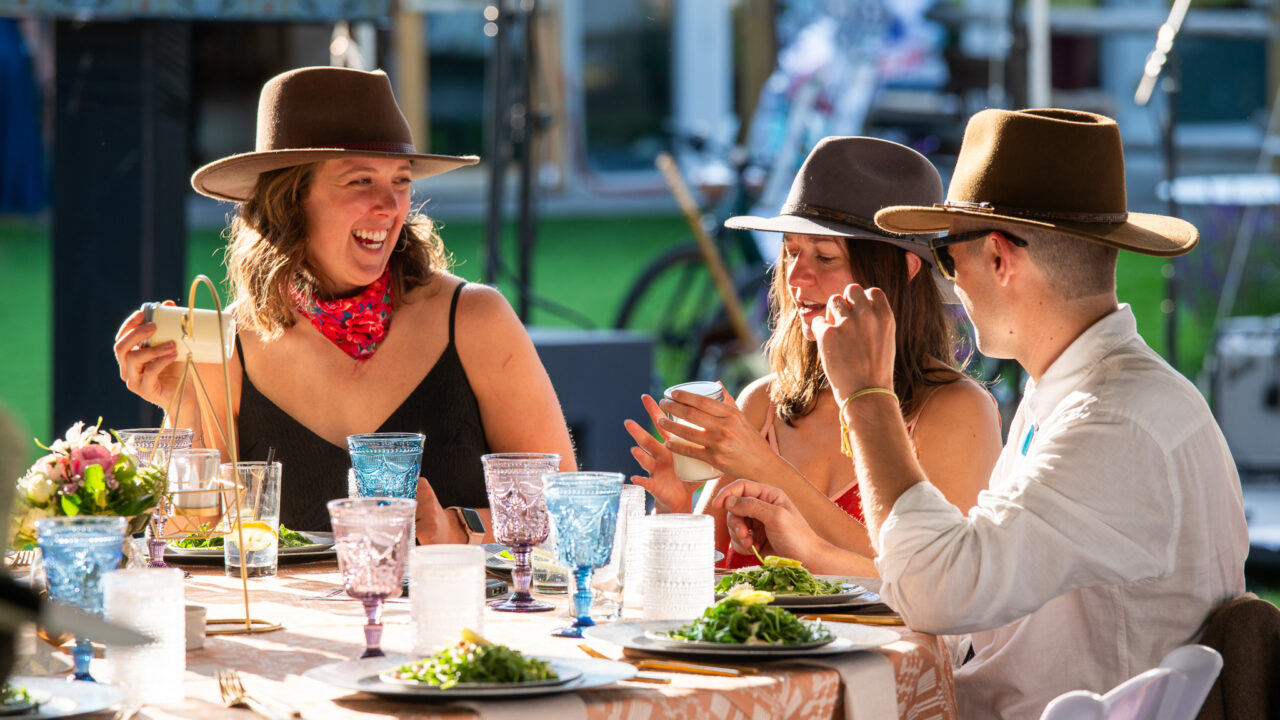 Three people in hats laughing at an outdoor table with food and drinks